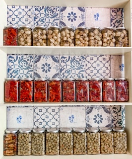 A wooden shelf displays three rows of glass jars filled with preserved food items. The top row contains jars of white mushrooms and artichokes, the middle row showcases jars of bright red peppers, and the bottom row contains jars of green olives. The background is adorned with white and blue patterned tiles, adding a traditional and decorative touch to the display.