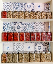 A wooden shelf displays three rows of glass jars filled with preserved food items. The top row contains jars of white mushrooms and artichokes, the middle row showcases jars of bright red peppers, and the bottom row contains jars of green olives. The background is adorned with white and blue patterned tiles, adding a traditional and decorative touch to the display.
