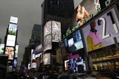 Bright electronic billboards and advertisements illuminate the bustling urban environment in an area crowded with vehicles and people. The signs include various brands, and the scene captures the vibrancy and energy of a commercial hub.