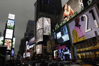 Bright electronic billboards and advertisements illuminate the bustling urban environment in an area crowded with vehicles and people. The signs include various brands, and the scene captures the vibrancy and energy of a commercial hub.