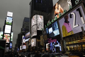 Bright electronic billboards and advertisements illuminate the bustling urban environment in an area crowded with vehicles and people. The signs include various brands, and the scene captures the vibrancy and energy of a commercial hub.