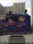 A large mural features a stylized illustration of a girl with long, dark hair and large eyes. She is set against a cosmic background with multicolored comets and planets. Below the mural, a planter displays the words 'IMAGINE PEACE' with greenery growing in it. The setting includes several tall buildings in an urban environment.