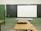 A classroom setting with a blackboard and a white projector screen at the front. There are wooden desks and chairs arranged in rows, facing the board. The walls are painted green, and the tiled floor is light-colored.