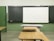 A classroom setting with a blackboard and a white projector screen at the front. There are wooden desks and chairs arranged in rows, facing the board. The walls are painted green, and the tiled floor is light-colored.
