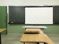 A classroom setting with a blackboard and a white projector screen at the front. There are wooden desks and chairs arranged in rows, facing the board. The walls are painted green, and the tiled floor is light-colored.