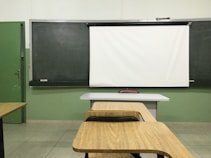A classroom setting with a blackboard and a white projector screen at the front. There are wooden desks and chairs arranged in rows, facing the board. The walls are painted green, and the tiled floor is light-colored.