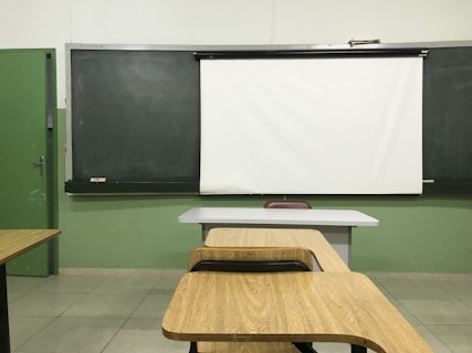A classroom setting with a blackboard and a white projector screen at the front. There are wooden desks and chairs arranged in rows, facing the board. The walls are painted green, and the tiled floor is light-colored.