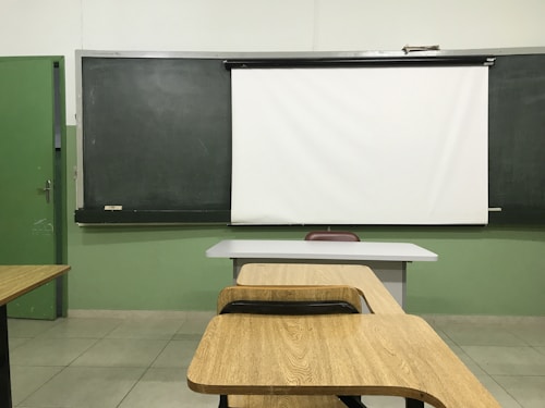 A classroom setting with a blackboard and a white projector screen at the front. There are wooden desks and chairs arranged in rows, facing the board. The walls are painted green, and the tiled floor is light-colored.
