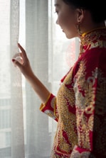 A person wearing an intricately embroidered red and gold traditional outfit stands by a window, gently touching a sheer curtain. The clothing features detailed patterns with visible gold and silver thread embellishments. The person's face is partially visible in profile, and earrings can be seen.