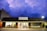 A store with a dark shingled roof and illuminated interior is set against a dramatic, cloudy sky at dusk. The sign reads 'Out of the Box - Games, Toys, Puzzles'. Inside, several people are seated at tables, possibly engaged in games or discussions.