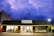 A store with a dark shingled roof and illuminated interior is set against a dramatic, cloudy sky at dusk. The sign reads 'Out of the Box - Games, Toys, Puzzles'. Inside, several people are seated at tables, possibly engaged in games or discussions.
