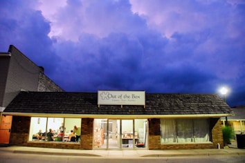 A store with a dark shingled roof and illuminated interior is set against a dramatic, cloudy sky at dusk. The sign reads 'Out of the Box - Games, Toys, Puzzles'. Inside, several people are seated at tables, possibly engaged in games or discussions.