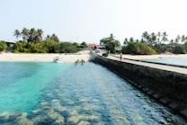 A serene beach scene with clear turquoise waters and a long pier leading towards a sandy shore. Lush greenery and palm trees line the background, offering a tropical feel. A small boat is anchored near the shore, and a person can be seen walking along the pier. The bright sunlight enhances the vibrant colors of the landscape.