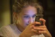 A young person is focused intently on a smartphone, holding it with both hands. The lighting creates a warm, intimate atmosphere with the screen's glow reflecting on their face.