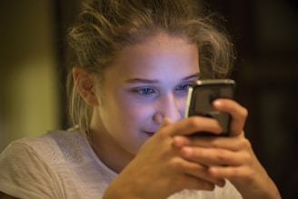 A young person is focused intently on a smartphone, holding it with both hands. The lighting creates a warm, intimate atmosphere with the screen's glow reflecting on their face.