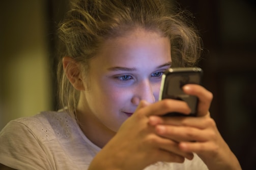 A young person is focused intently on a smartphone, holding it with both hands. The lighting creates a warm, intimate atmosphere with the screen's glow reflecting on their face.