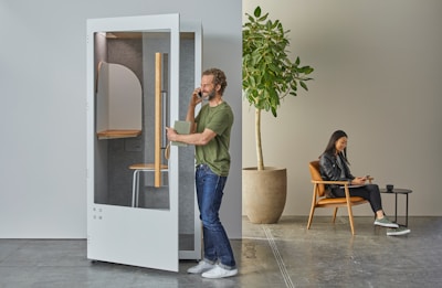 A man is using a phone booth for a call, holding a notebook in one hand. To the right, a woman is sitting in a modern chair using a tablet. A potted plant is nearby, adding a natural element to the minimalist setting.