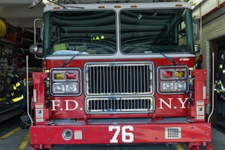 A red fire truck parked in a garage with the letters F.D. and N.Y. visible on its front. The number 76 is prominently displayed below the grille. Various firefighting equipment such as hoses and gear is visible in the background.
