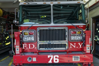 A red fire truck parked in a garage with the letters F.D. and N.Y. visible on its front. The number 76 is prominently displayed below the grille. Various firefighting equipment such as hoses and gear is visible in the background.