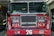 A red fire truck parked in a garage with the letters F.D. and N.Y. visible on its front. The number 76 is prominently displayed below the grille. Various firefighting equipment such as hoses and gear is visible in the background.