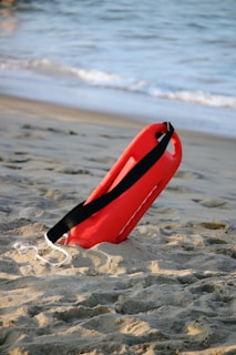 A red lifeguard float with a black strap is positioned upright in the sand, set against the backdrop of a beach with gentle ocean waves.