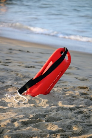 A red lifeguard float with a black strap is positioned upright in the sand, set against the backdrop of a beach with gentle ocean waves.