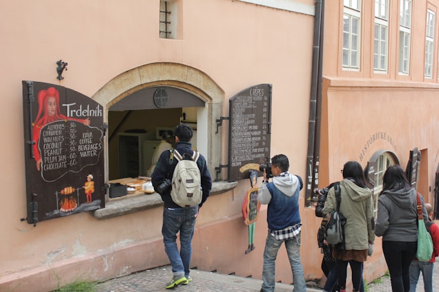 A small food stall built into a wall offers traditional pastries called Trdelník. Several people are gathered around, capturing photos and looking at the menu, which lists various flavor options. The stall has a rustic appearance, with wooden boards displaying menu items.