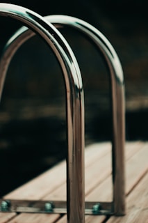 Two metallic, polished handrails are installed on a wooden deck, likely part of a swimming pool area. The handrails are shiny and create a sleek, modern aesthetic against the blurred dark background.