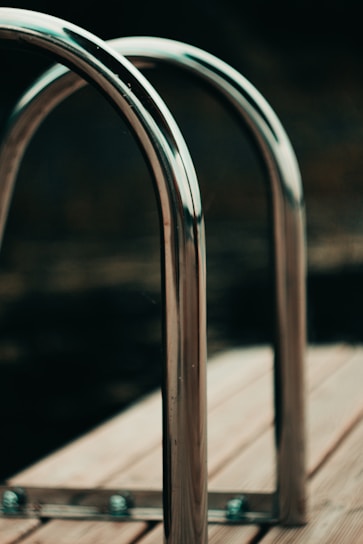 Two metallic, polished handrails are installed on a wooden deck, likely part of a swimming pool area. The handrails are shiny and create a sleek, modern aesthetic against the blurred dark background.