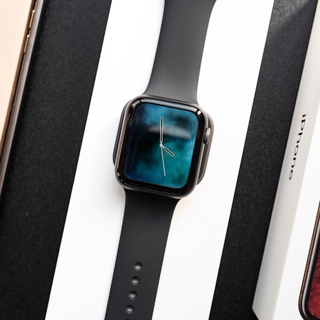 A black smartwatch with a digital clock display showing hands on a gradient blue and black background. It is placed on a white surface between a boxed phone labeled 'iPhone' and another boxed item with a visible wooden texture.