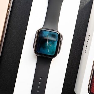 A black smartwatch with a digital clock display showing hands on a gradient blue and black background. It is placed on a white surface between a boxed phone labeled 'iPhone' and another boxed item with a visible wooden texture.