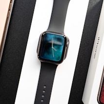A black smartwatch with a digital clock display showing hands on a gradient blue and black background. It is placed on a white surface between a boxed phone labeled 'iPhone' and another boxed item with a visible wooden texture.