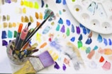 A collection of art supplies including various paintbrushes and markers is placed in a holder. The background features a palette and a sheet with numerous colorful paint swatches. The palette has multiple sections for mixing colors.