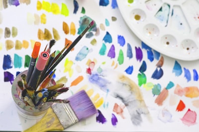 A collection of art supplies including various paintbrushes and markers is placed in a holder. The background features a palette and a sheet with numerous colorful paint swatches. The palette has multiple sections for mixing colors.