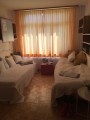 A cozy room with two single beds arranged parallel to each other, both adorned with striped pillows and light blankets. The large window is covered by light peach curtains that diffuse the sunlight, creating a warm glow. A red bedside table holding a picture frame is situated between the beds. Wooden flooring and shelves add to the room's cozy feel, and ski boots are placed near a white round side table with a plant on top.