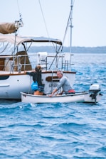 Two people are in a small motorized dinghy, approaching a larger sailboat. One person is leaning towards the larger boat, attempting to climb onboard, while the other is steering the dinghy. The surrounding water is a vibrant blue with gentle waves, and the sky appears calm, indicating a peaceful day at sea.