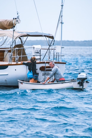 Two people are in a small motorized dinghy, approaching a larger sailboat. One person is leaning towards the larger boat, attempting to climb onboard, while the other is steering the dinghy. The surrounding water is a vibrant blue with gentle waves, and the sky appears calm, indicating a peaceful day at sea.