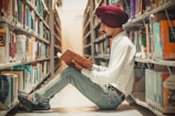 A person wearing a maroon turban and glasses is sitting on the floor of a library aisle, surrounded by bookshelves filled with books. They are reading an open book, holding it with both hands. The environment conveys a quiet and studious atmosphere.