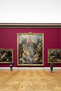 An art gallery with a large, ornate painting mounted on a deep red wall. The painting features numerous figures in dynamic poses, surrounded by intricate details. Two smaller paintings are on either side. Two visitors are observing the artwork; one is using crutches.