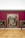 An art gallery with a large, ornate painting mounted on a deep red wall. The painting features numerous figures in dynamic poses, surrounded by intricate details. Two smaller paintings are on either side. Two visitors are observing the artwork; one is using crutches.