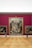 An art gallery with a large, ornate painting mounted on a deep red wall. The painting features numerous figures in dynamic poses, surrounded by intricate details. Two smaller paintings are on either side. Two visitors are observing the artwork; one is using crutches.