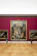 An art gallery with a large, ornate painting mounted on a deep red wall. The painting features numerous figures in dynamic poses, surrounded by intricate details. Two smaller paintings are on either side. Two visitors are observing the artwork; one is using crutches.