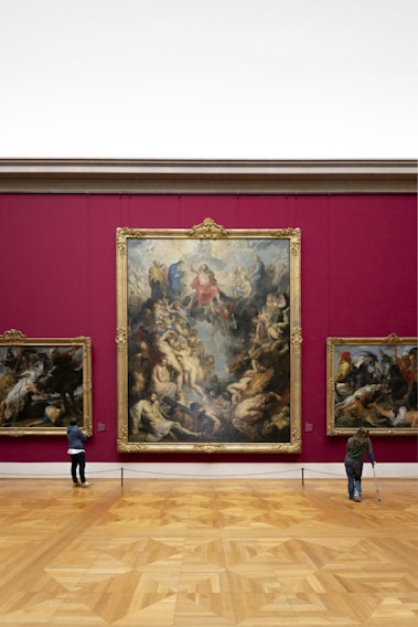 An art gallery with a large, ornate painting mounted on a deep red wall. The painting features numerous figures in dynamic poses, surrounded by intricate details. Two smaller paintings are on either side. Two visitors are observing the artwork; one is using crutches.