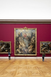 An art gallery with a large, ornate painting mounted on a deep red wall. The painting features numerous figures in dynamic poses, surrounded by intricate details. Two smaller paintings are on either side. Two visitors are observing the artwork; one is using crutches.