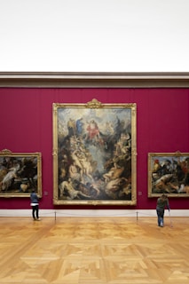 An art gallery with a large, ornate painting mounted on a deep red wall. The painting features numerous figures in dynamic poses, surrounded by intricate details. Two smaller paintings are on either side. Two visitors are observing the artwork; one is using crutches.