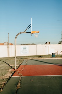 A basketball hoop stands on an outdoor court with a red and green surface. The court is surrounded by a low white wall with grass bordering it. The sky is clear and blue, and there is a trash can to the side.