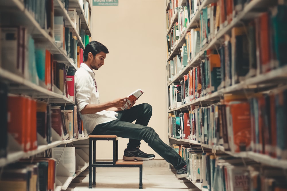 man sitting and leaning near bookshelf while reading book