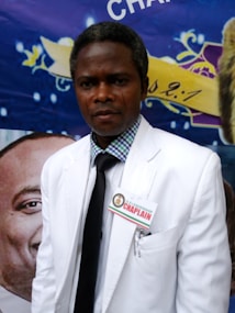 A person wearing a white suit and a name tag labeled 'Chaplain' is standing in front of a colorful backdrop. The backdrop features vibrant colors and decorative elements, including a partial view of a lion emblem and some text.