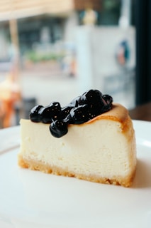 A slice of creamy cheesecake with a golden-brown crust, topped with a generous serving of dark blueberry sauce, placed on a white plate. The background is softly blurred, indicating an indoor setting with large windows.