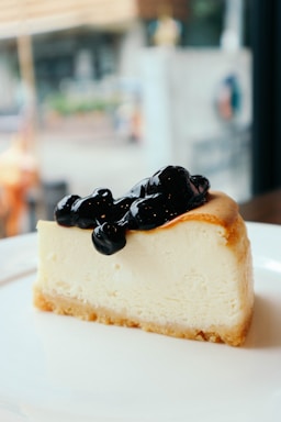 A slice of creamy cheesecake with a golden-brown crust, topped with a generous serving of dark blueberry sauce, placed on a white plate. The background is softly blurred, indicating an indoor setting with large windows.